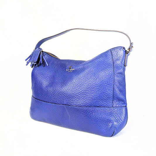 A blue pebbled leather Kate Spade handbag with a tassel, showcasing its side profile with a visible logo and gold-tone hardware.